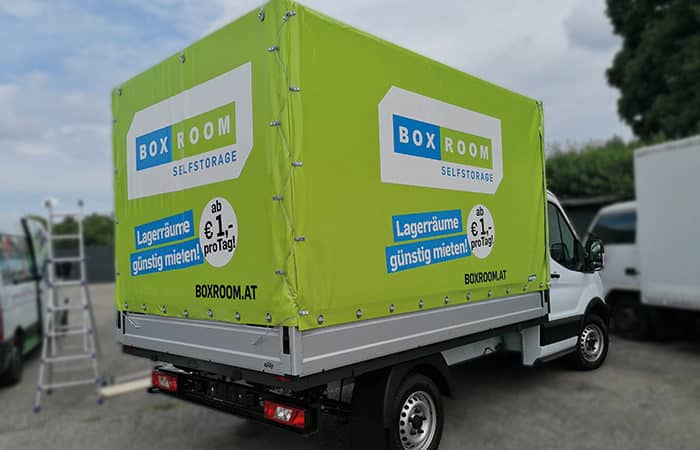 Das Bild zeigt ein Pritschenfahrzeug der Firma Boxroom Selfstorage mit einer Vollplane und einem beeindruckenden Digitaldruck. Die Plane schützt die Ladung vor Witterungseinflüssen und bietet eine effektive Werbefläche für das Unternehmen. Der Transporter ermöglicht es Boxroom Selfstorage, ihre Dienstleistungen auf effektive Weise zu bewerben und gleichzeitig Güter sicher und geschützt zu transportieren.