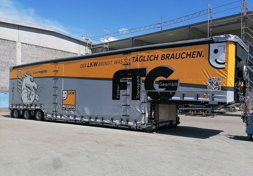 Das Bild zeigt einen modernen und professionellen Meusburger Auflieger der Firma FTG, der nicht nur für den Transport von Maschinen, sondern auch als mobile Werbeplattform genutzt wird.